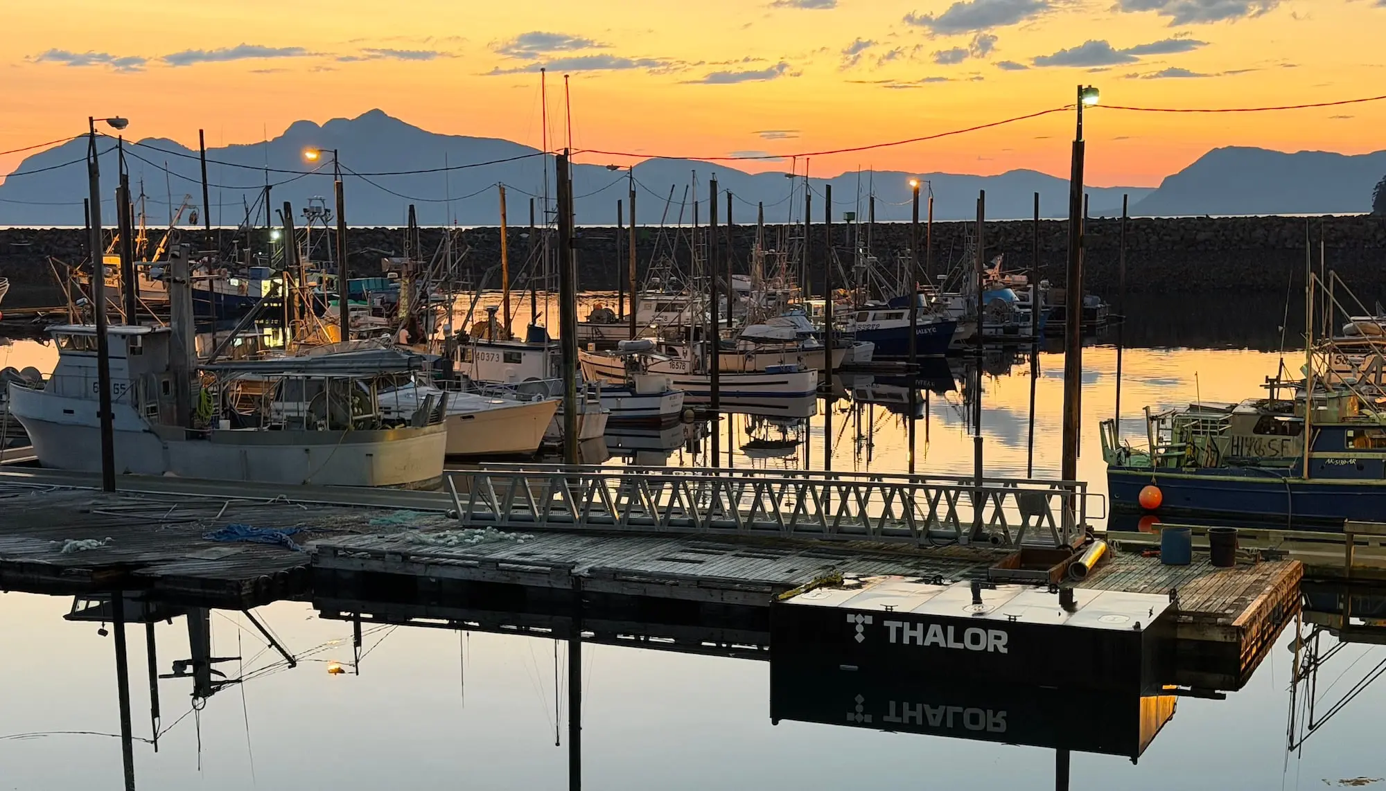 A Thalor work platform moored at the Metlakatla harbor float at sunset, fishing boats behind
